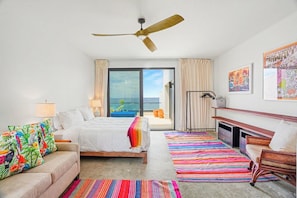 Habitación de hotel con cama, sofá, televisión, ventilador de techo y balcón con vista al océano.