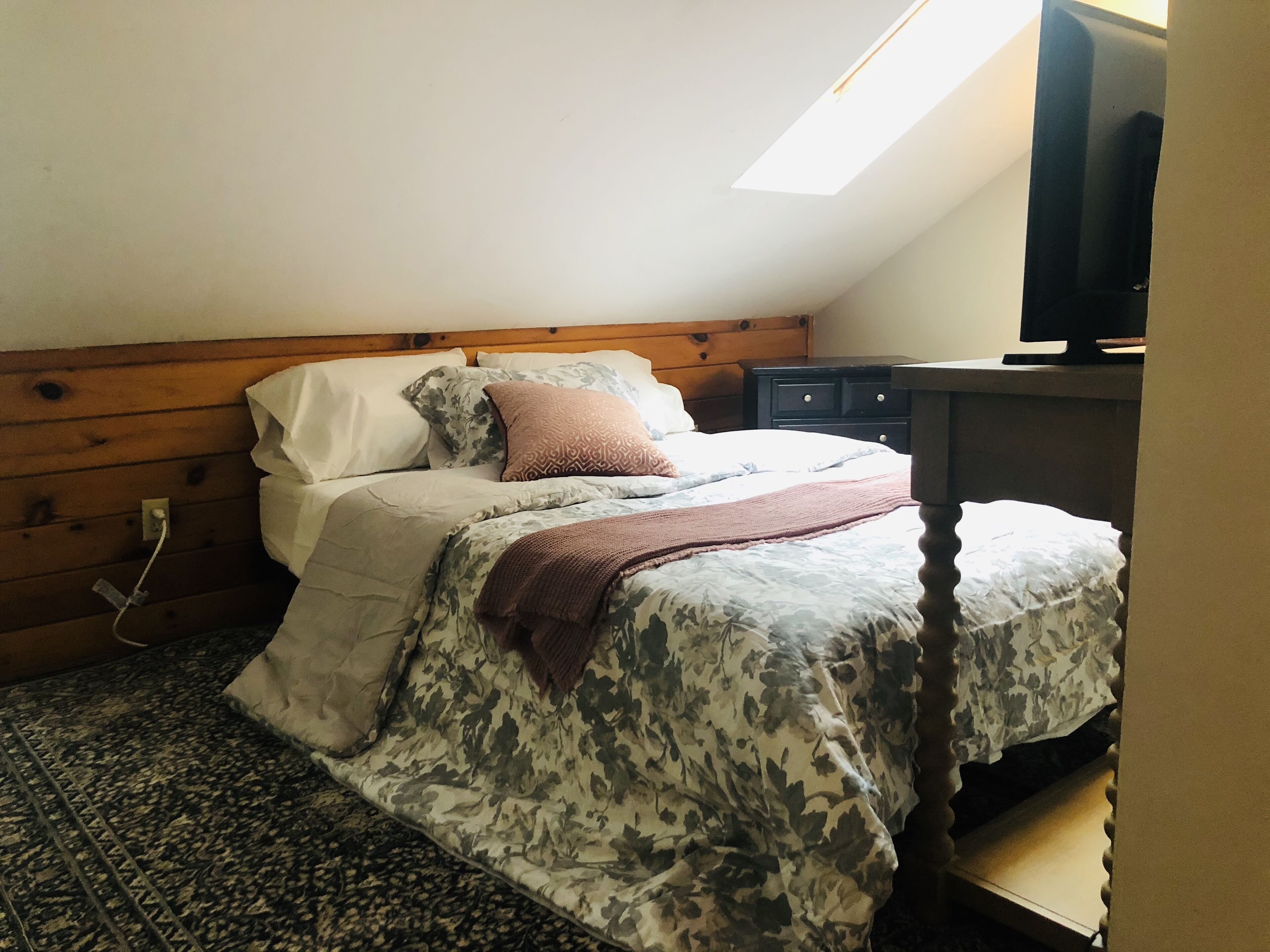 A bedroom with a bed, wooden headboard, a small desk, and a television on a stand.