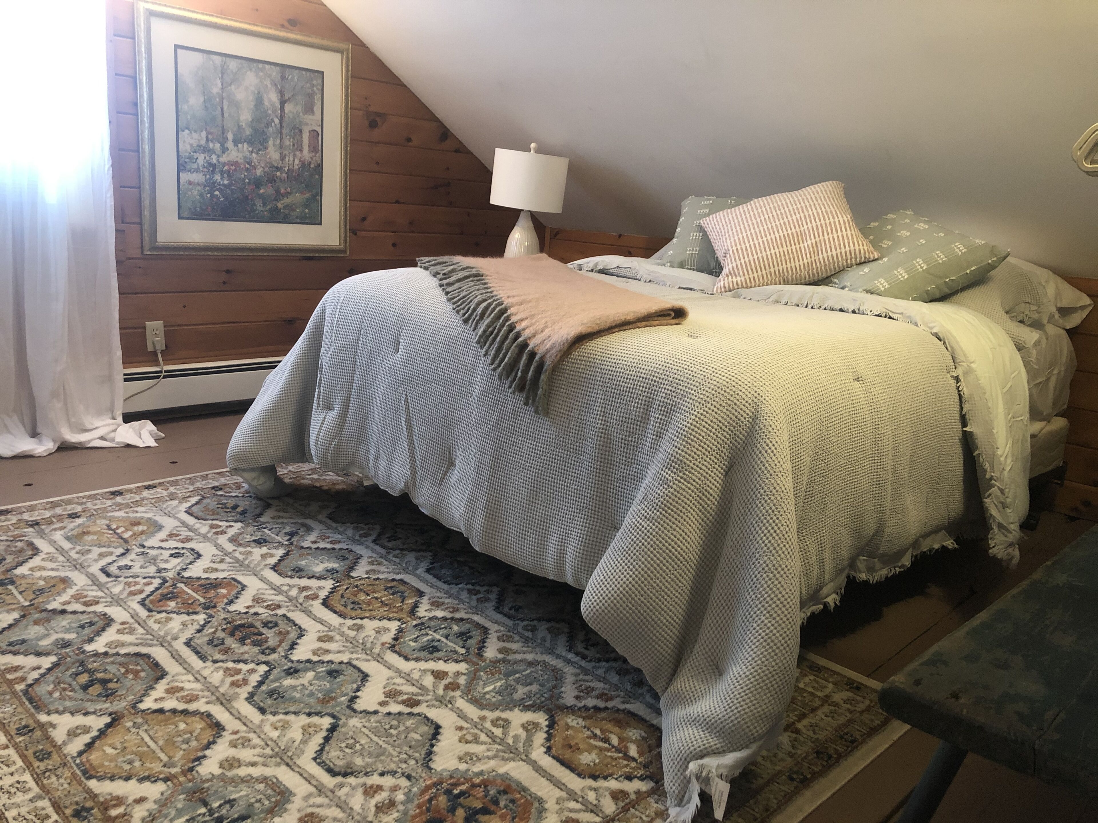 A cozy bedroom with a bed, pillows, a lamp, and a patterned rug.