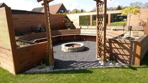 Une terrasse en bois dotée d’un foyer circulaire, entourée de parterres surélevés et d’une zone recouverte de gravier.