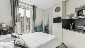 A hotel room with a bed, a desk, a chair, a TV, and a view of buildings through the window. - Studio Mac Mahon - Champs Elysees by B'Your Home (Paris)