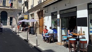 Eine Straße mit Außenbestuhlung vor einem Café, ein Bankschild und ein Gebäude im klassischen Stil.