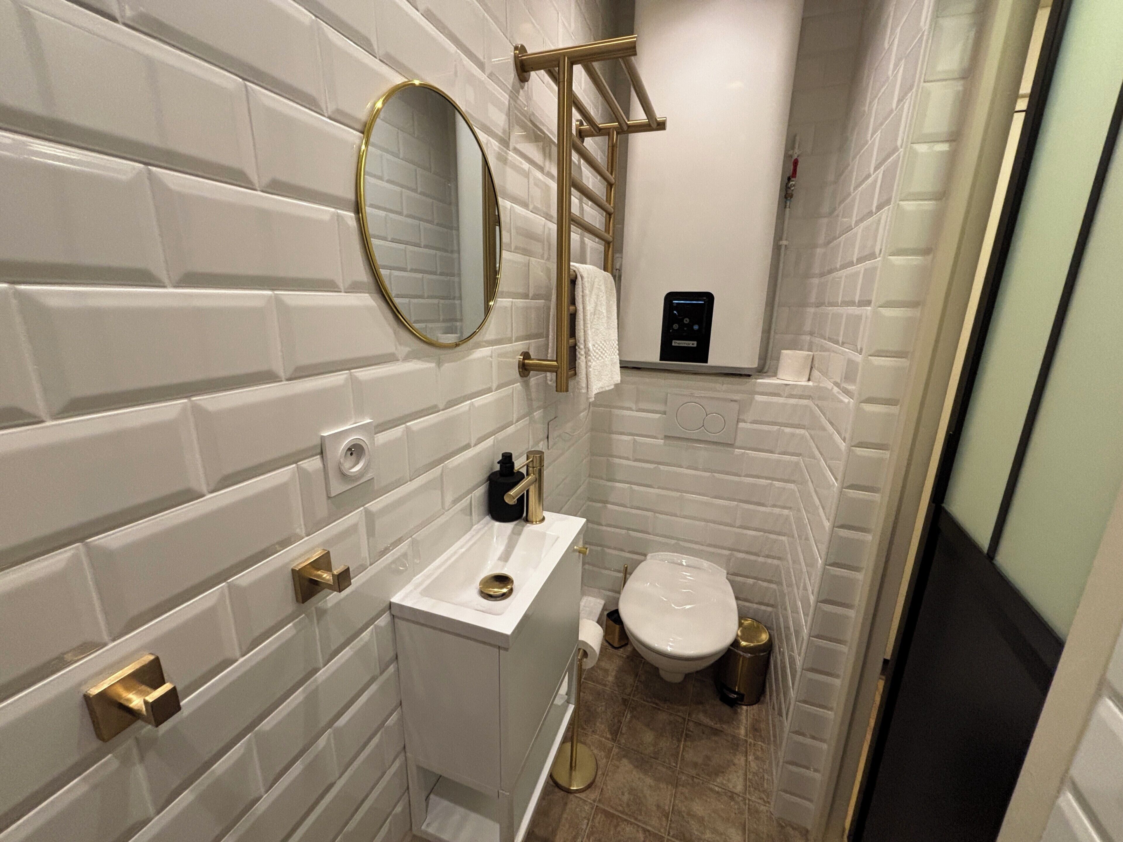 A compact bathroom with white subway tile walls, a gold-framed mirror, a white sink with a gold faucet, a toilet, and a trash can.