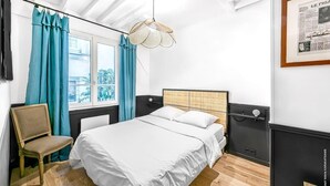Ein Schlafzimmer mit einem Bett, einem Sessel, einem Fenster mit Vorhängen, einer Pendelleuchte und einem gerahmten Bild an der Wand.