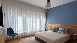 A bedroom with a large bed, a chair, a lamp, and sheer curtains. - Oceanus Executive Apartamento (Ponta Delgada)