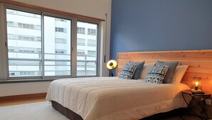 A bedroom with a large bed, a nightstand with a lamp, a window with a view of buildings, and a blue wall. - Oceanus Executive Apartamento (Ponta Delgada)