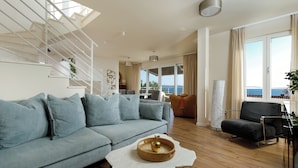 Ein modernes Wohnzimmer mit einem grauen Sofa, einem schwarzen Sessel und einem Holzboden. Eine Treppe mit weißen Geländern und Blick auf das Meer durch große Fenster.