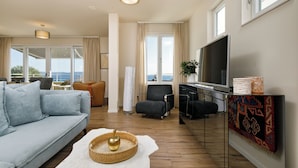 Ein modernes Wohnzimmer mit Sofa, Sesseln, Fernseher und Blick ins Freie.
