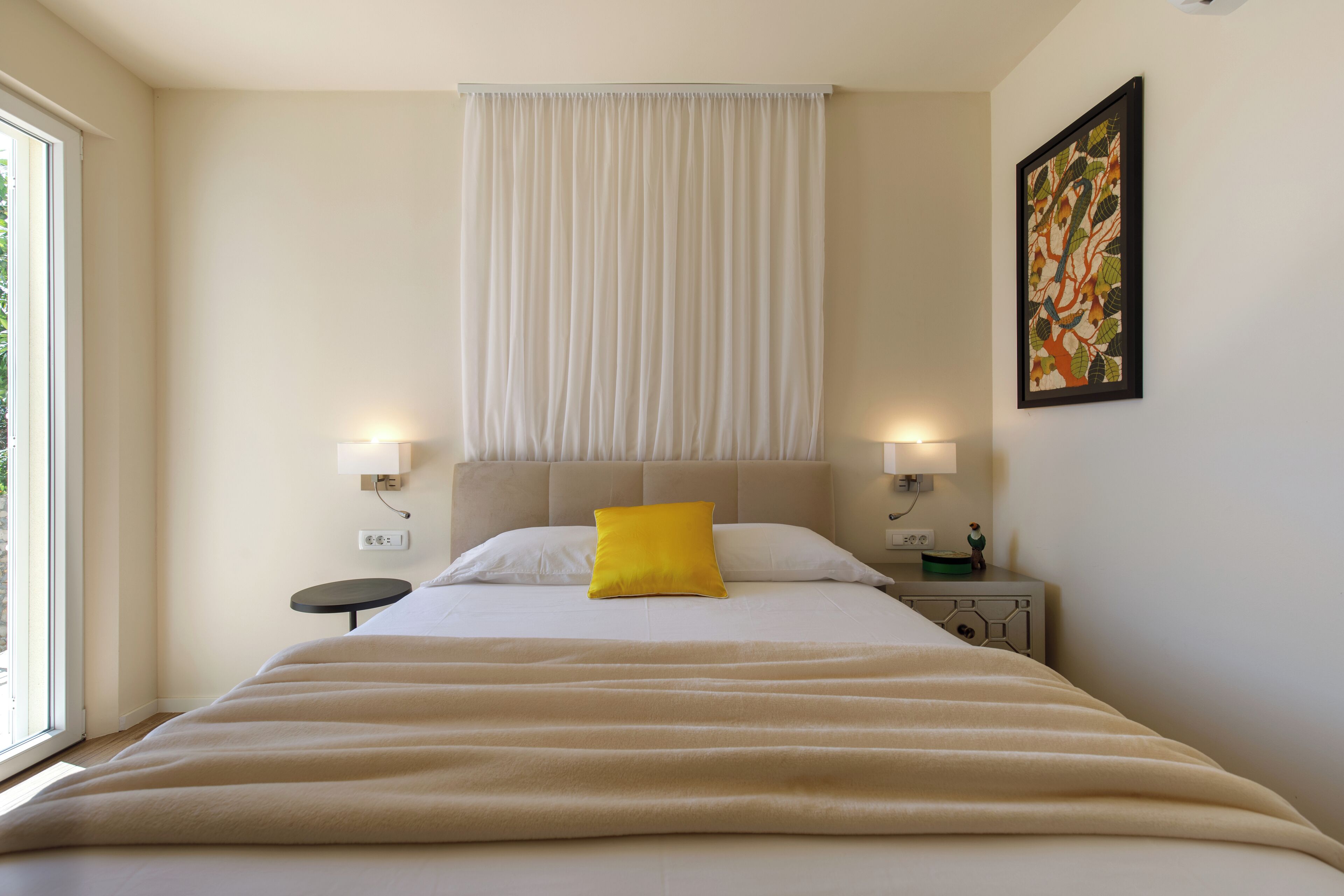 A hotel room with a large bed, a bedside table, a nightstand, a window, and a wall-mounted painting.