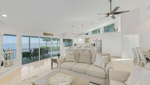 A spacious living room with a large window overlooking a body of water, a ceiling fan, and a kitchen area visible through the glass doors. - Bay beauty with heated pool, spa, games, ping pong (Holmes Beach)
