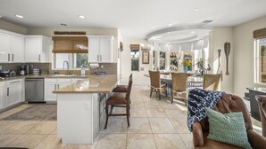 A spacious kitchen with white cabinets, a granite countertop, and a central island. There is a dining area with wicker chairs and a table. - 🏝️Chic villa on the lake! 3 bed 3 bath, Golf, bbq & fun in the sun 🌵🏝️🍦👙 (Indio)