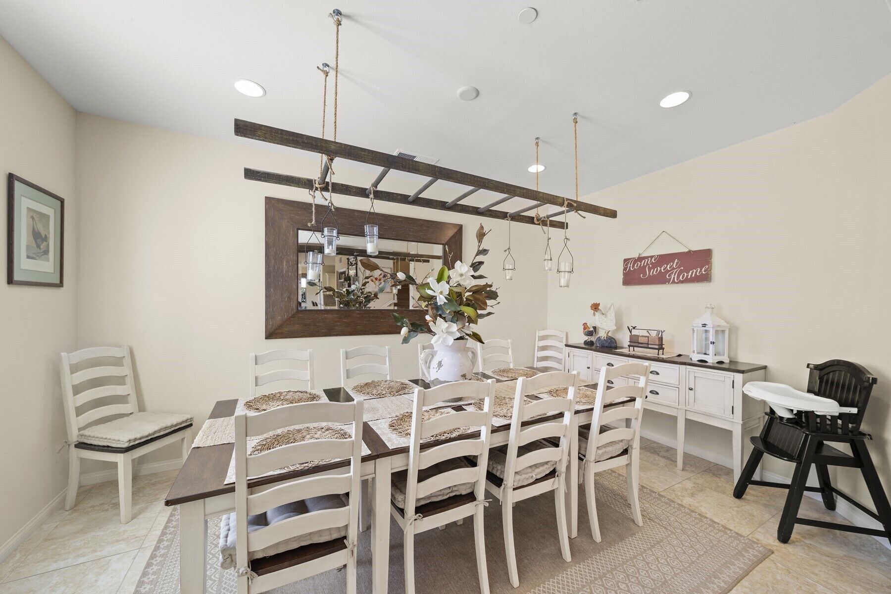 Un coin repas avec une table en bois et des chaises blanches, un miroir, des luminaires suspendus et une pancarte indiquant « Chez soi, quel bonheur ! ».