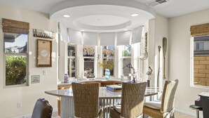A dining area with a round table, wicker chairs, and a view of the outdoors through large windows. - 🏝️Chic villa on the lake! 3 bed 3 bath, Golf, bbq & fun in the sun 🌵🏝️🍦👙 (Indio)