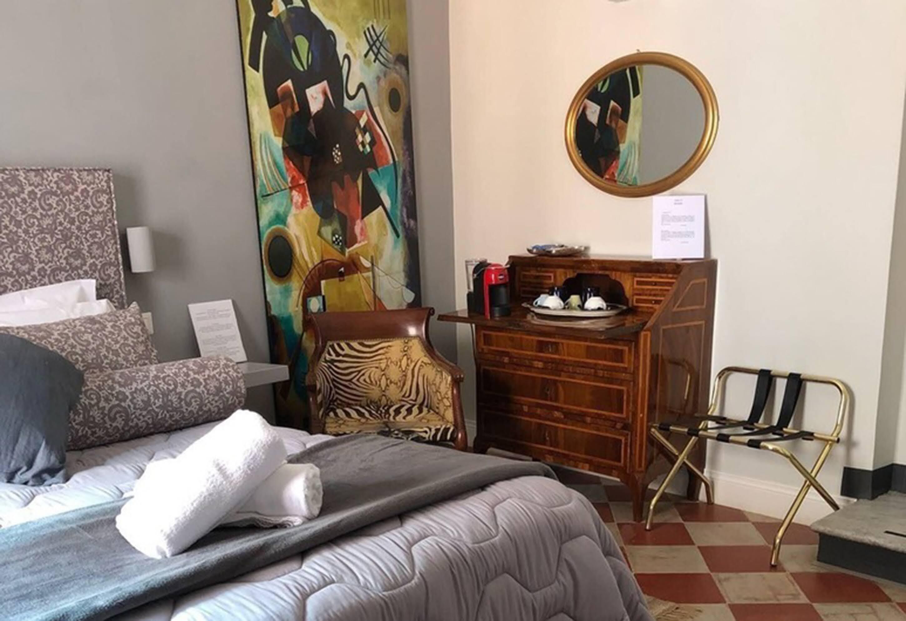 Habitación de hotel con cama, cómoda, silla de estampado de cebra, espejo y un cuadro abstracto de colores en la pared.