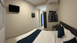 A hotel room with a bed, a mounted TV, an air conditioning unit, a door, and a wall-mounted light.