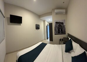 A hotel room with a bed, a mounted TV, an air conditioning unit, a door, and a wall-mounted light.