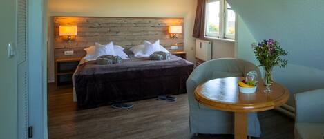 Ein Hotelzimmer mit einem hölzernen Kopfteil, einem Bett mit weißer Bettwäsche, einem runden Holztisch mit einer Vase und Obst sowie einem Stuhl.