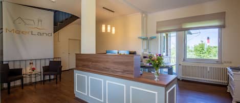 A reception area with a wooden reception desk, a seating area with chairs, a staircase, pendant lights, and a window with a view of greenery.