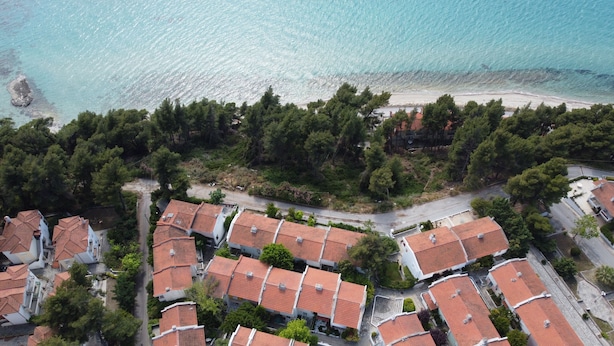 An aerial view of a coastal residential area with terraced houses, a beach, and dense greenery. - Zennova 16 Naftilos Family Summer House (Kassandra)
