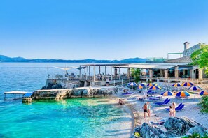 A coastal hotel with a pier extending into the sea, beach umbrellas, and lounge chairs.