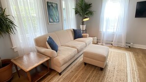 A living room with a beige sofa, a matching ottoman, a small wooden side table, and a large woven rug. - Charming, Central Jamestown Cottage, walk to town/beach! (Jamestown)