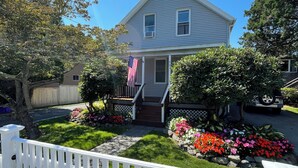 A two-story house with a front porch, a small garden with colorful flowers, and a white picket fence. - Charming, Central Jamestown Cottage, walk to town/beach! (Jamestown)