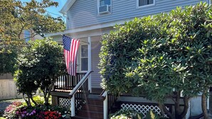 A two-story house with a front porch, a flag, and a well-maintained garden with colorful flowers and shrubs. - Charming, Central Jamestown Cottage, walk to town/beach! (Jamestown)