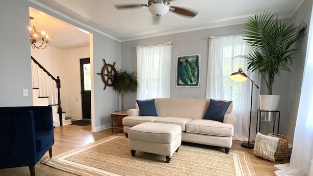 A living room with a beige sofa, a blue armchair, a rug, a ceiling fan, a plant, and a painting on the wall. - Charming, Central Jamestown Cottage, walk to town/beach! (Jamestown)