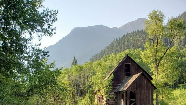A rustic wooden structure built on a rock ledge by a flowing river, surrounded by lush greenery and mountains in the background.