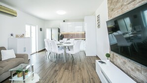 A modern living room with a dining area, a kitchen in the background, and a large flat-screen TV mounted on the wall. - Modern House W.large Garden & Children Playground (Split)