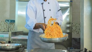 A chef in a professional kitchen holding a large metal bowl of spaghetti.