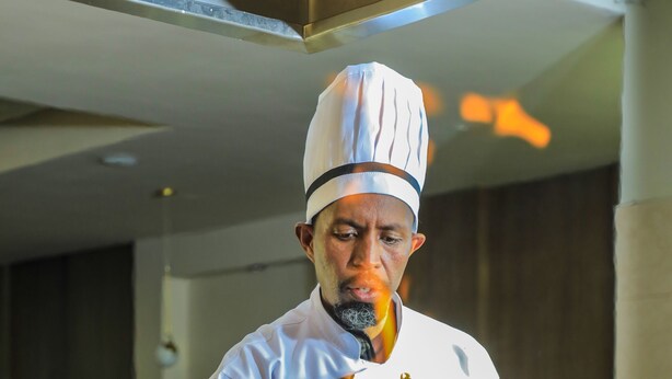 A chef in a white uniform with a tall hat, standing in a kitchen with a grill and flames.