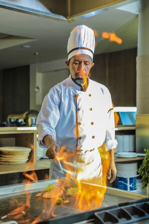 Ein Koch in einer weißen Uniform mit Kochhut steht in einer Küche mit Grill und Flammen.