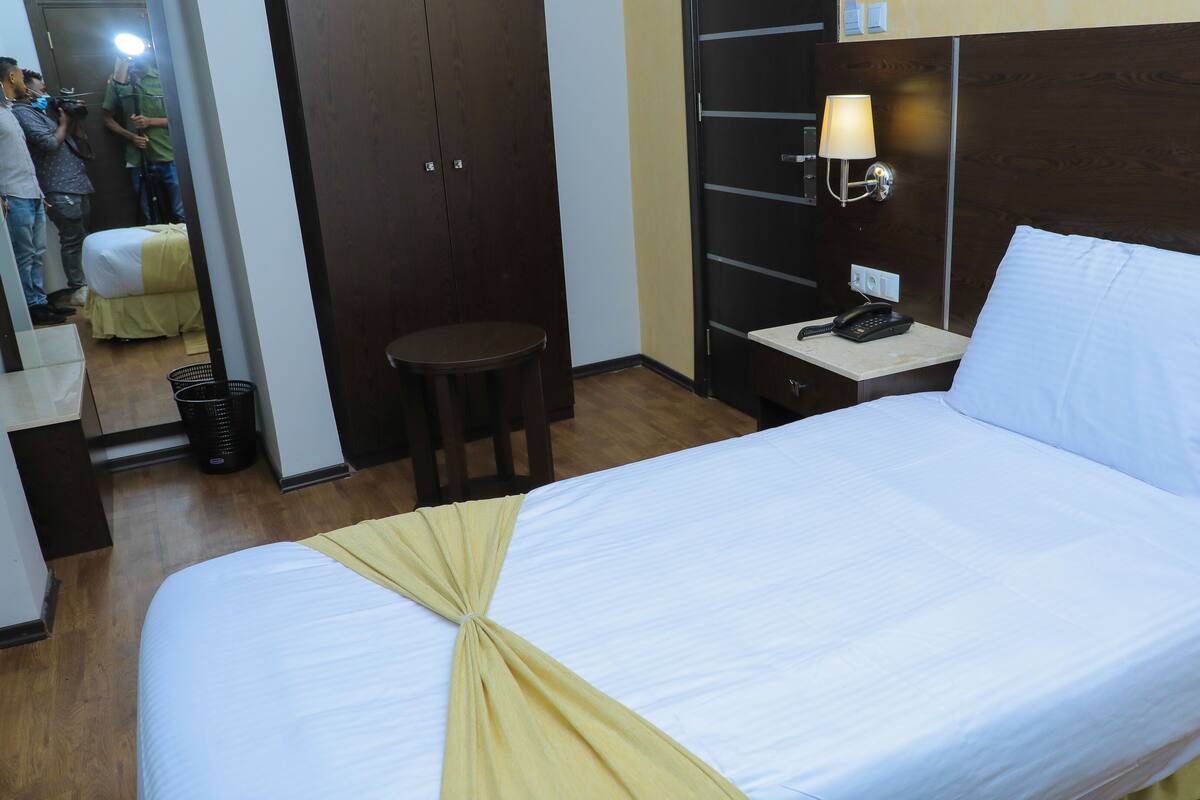 A hotel room with a bed, a nightstand with a lamp, a mirror, a small stool, and a wooden wardrobe.