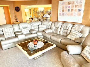 A modern living room with a large L-shaped sectional sofa, a coffee table with decorative items, a wall clock, and a framed artwork. - Maisons Sur Mer -Oceanfront Luxury (Myrtle Beach)