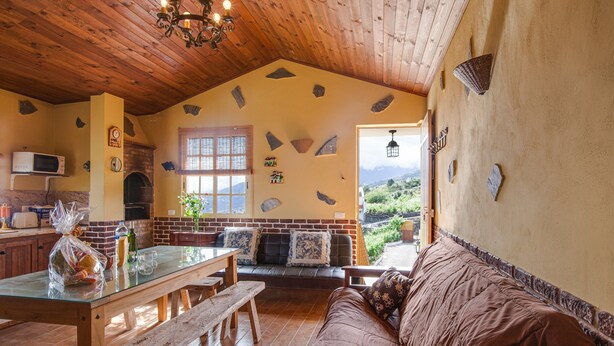 A cozy interior with a wooden ceiling, a stone wall, a glass table, a brown sofa, and a view of greenery outside. - Vacation Home Casa La Chocita with Sea View, Mountain View (Los Realejos)