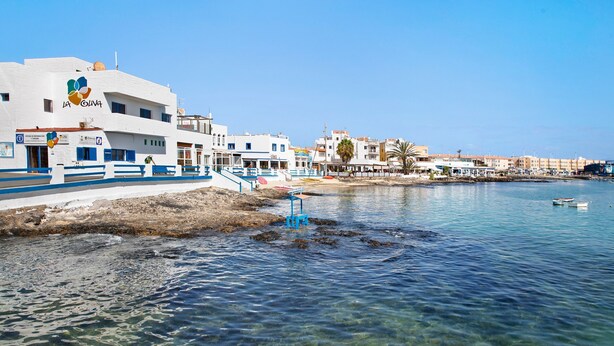 Un bâtiment situé sur la côte, offrant une vue dégagée sur la mer, un escalier bleu menant à la plage et un petit bateau sur l’eau.