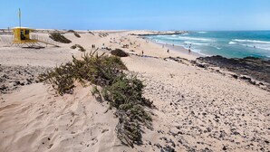 Une plage de sable avec une tour de surveillance pour les sauveteurs, une côte rocheuse et une végétation clairsemée.