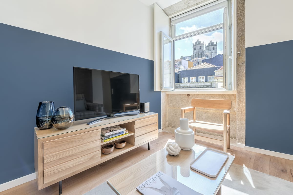 Une chambre moderne dotée d’une grande fenêtre, d’un téléviseur à écran plat, d’une console en bois et offrant une vue sur un bâtiment historique.