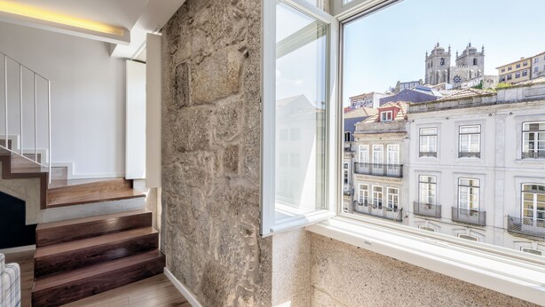 Un intérieur moderne avec un mur en pierre, un escalier en bois et une grande fenêtre donnant sur des bâtiments historiques.