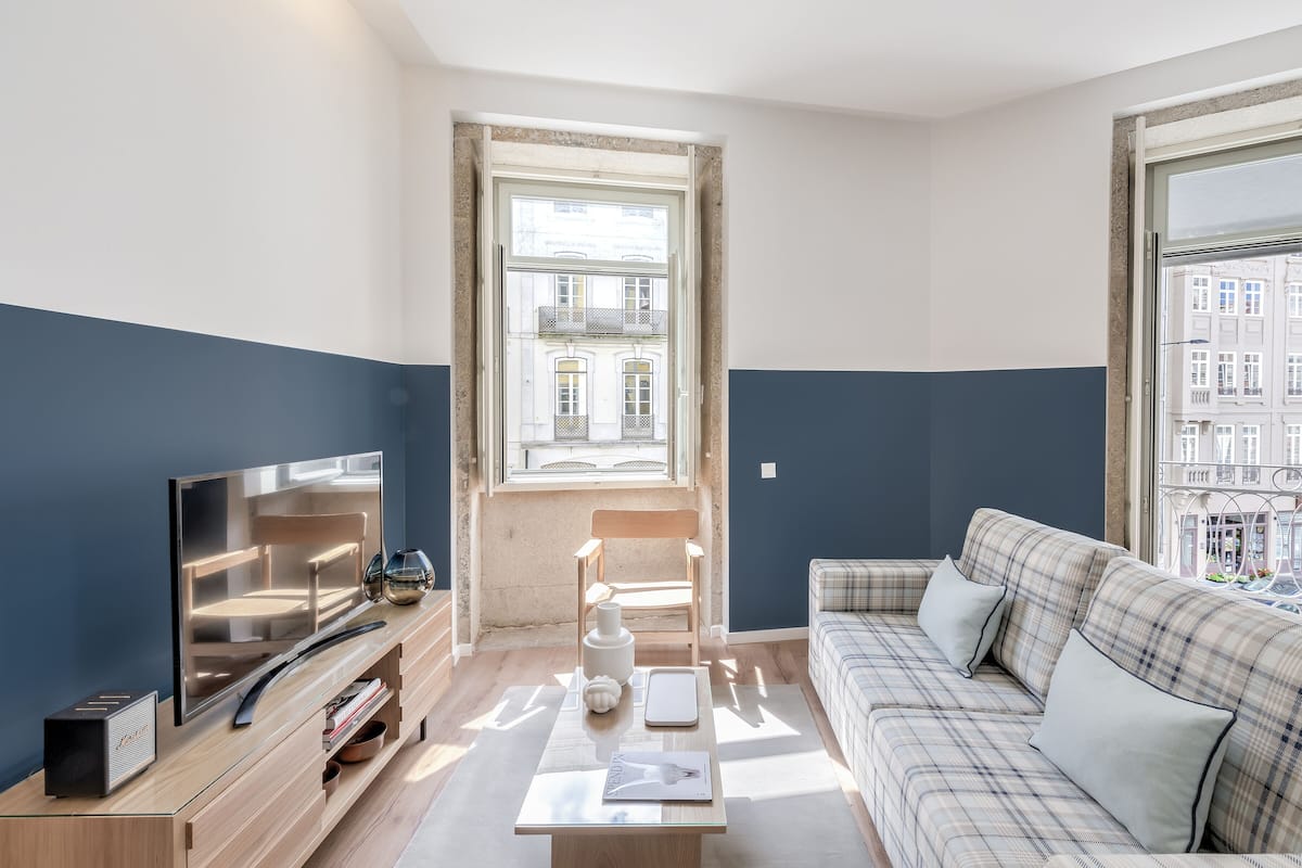 Un salon moderne avec un téléviseur à écran plat, une table basse en bois, un canapé à motifs écossais et une vue sur les immeubles par la fenêtre.