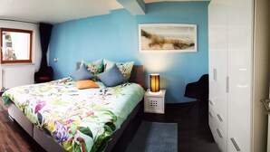 Une chambre à coucher avec une couvre-lit à motifs floraux, une table de chevet avec une lampe, une fenêtre avec des rideaux et une décoration murale sur le thème de la plage.