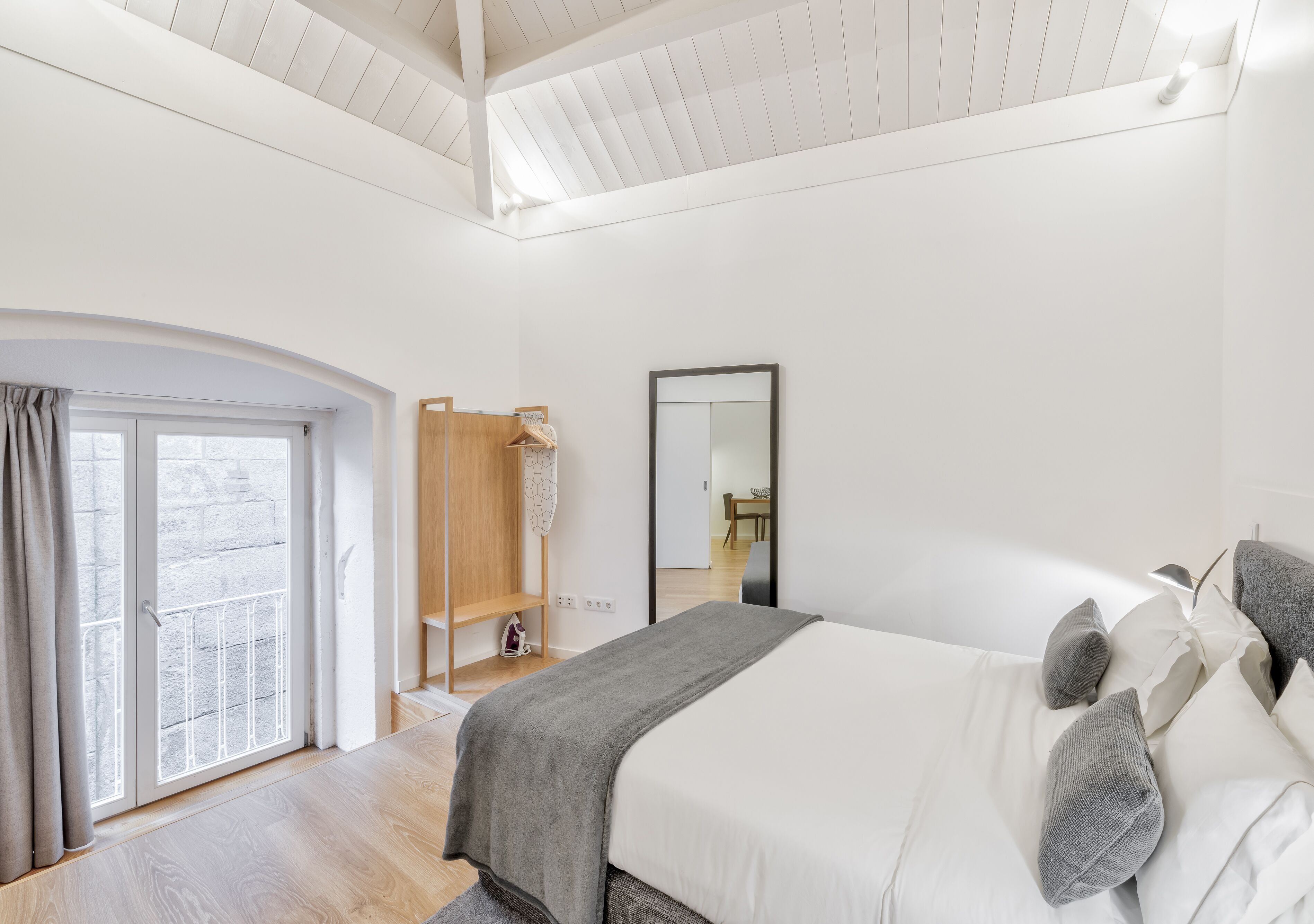 A modern bedroom with a large bed, a wooden wardrobe, a mirror, and a view of the outdoors through a glass door.
