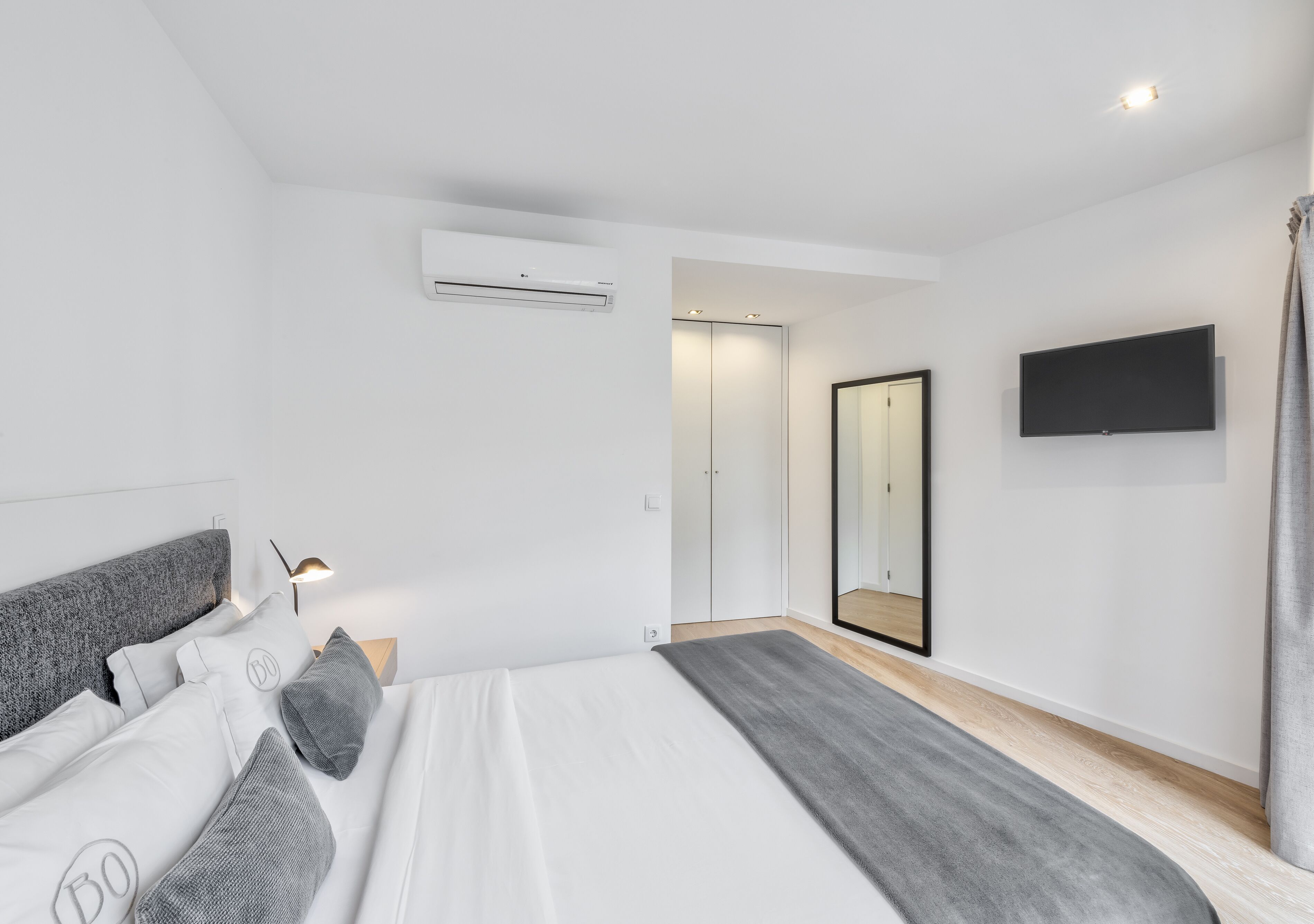 A modern bedroom with a bed, bedside table, lamp, air conditioning unit, wardrobe, mirror, and a TV mounted on the wall.