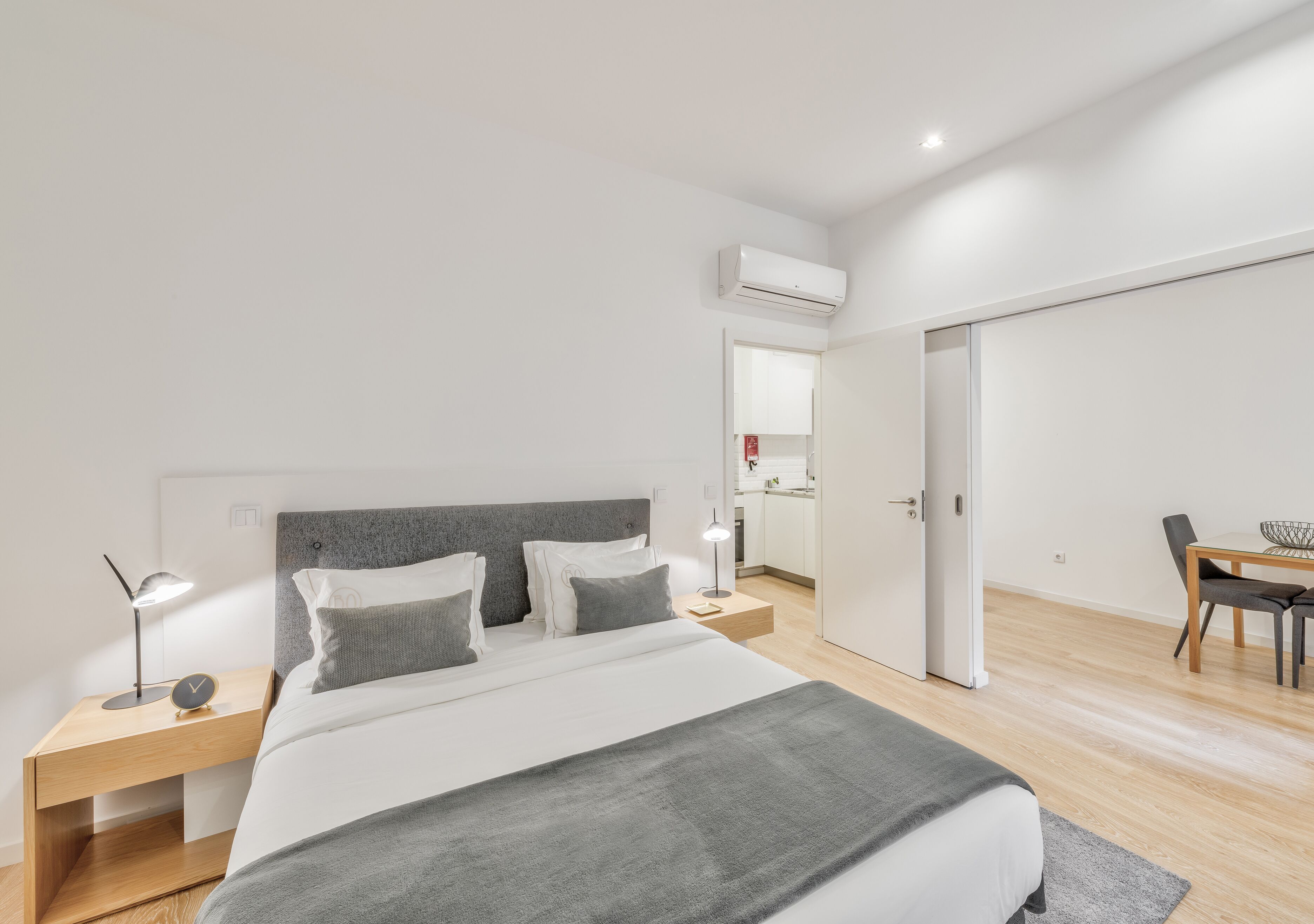 A modern bedroom with a large bed, bedside tables, a desk, and an open door leading to a kitchen.