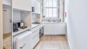 Eine kompakte Küche mit weißen Schränken, einem eingebauten Backofen, einer Kaffeemaschine und einem Fenster mit Blick auf die Straße.