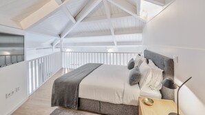 Ein modernes Schlafzimmer mit schräg versiegelter Decke, einem großen Bett mit grauem Kopfteil, einem Nachttisch mit Lampe und Blick auf einen Balkon.