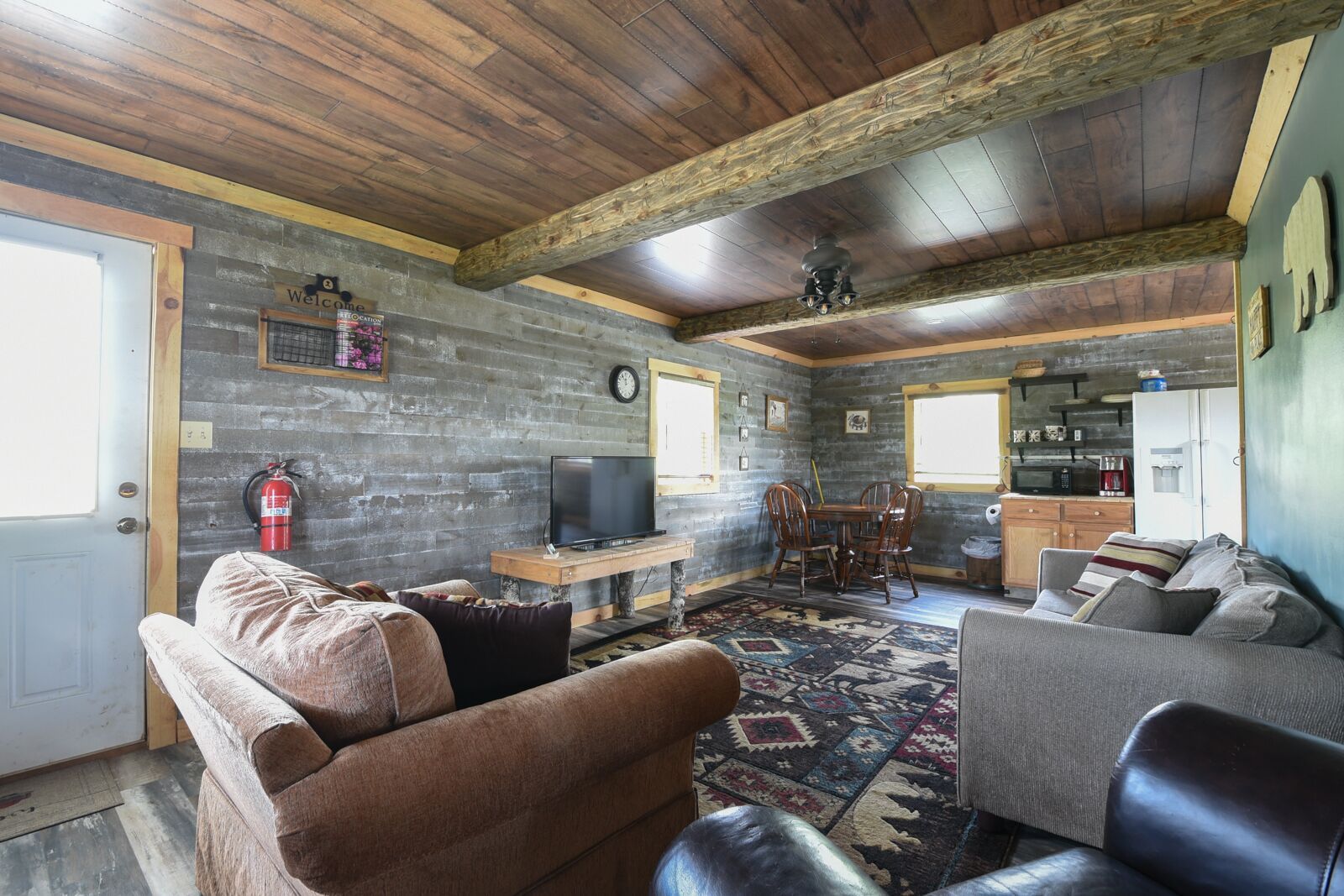 A rustic interior with a wooden ceiling, a television on a wooden stand, a dining table with chairs, a kitchen area with appliances, and a fire extinguisher on the wall.