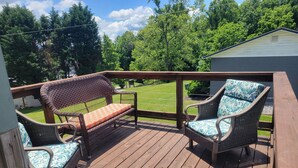 A wooden deck with wicker furniture and a view of a green lawn and trees. - Charming one room cabin perfect for couples or small families. (Flat Rock)