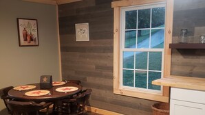 A dining area with a wooden table and chairs, a window with a view of a grassy path, and a kitchen area with a wooden counter and cabinets. - Charming one room cabin perfect for couples or small families. (Flat Rock)